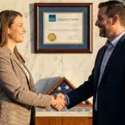 Two business professionals shaking hands in front of a framed GSA Multiple Award Schedule (MAS) contract certificate to celebrate a successful federal procurement award.