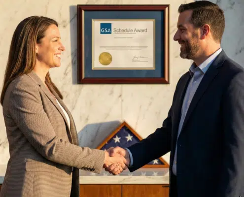 Two business professionals shaking hands in front of a framed GSA Multiple Award Schedule (MAS) contract certificate to celebrate a successful federal procurement award.