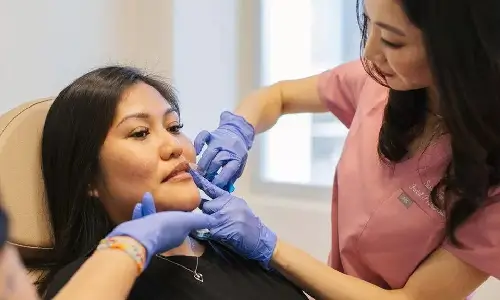 Injector, Dr Susan Tan, in pink scrubs and purple gloves placing dermal filler near a seated patient's lower lip during a preventive skin care visit.