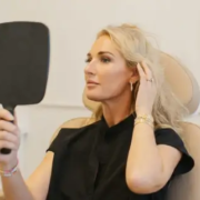 A blonde woman in a black shirt sits in a clinic chair looking into a hand mirror, portraying an executive refresh via subtle cosmetic surgery.