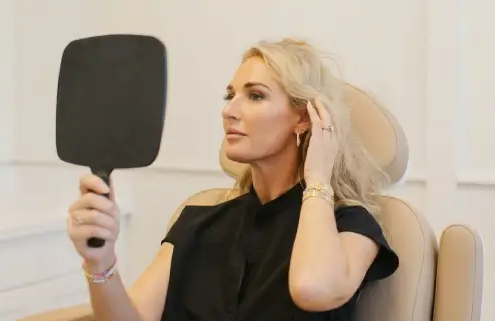 A blonde woman in a black shirt sits in a clinic chair looking into a hand mirror, portraying an executive refresh via subtle cosmetic surgery.