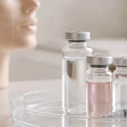 Dermal filler vials with silver caps and a marked syringe on a glass tray beside a mannequin head mapped with facial contour lines.